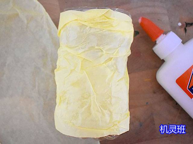 儿童用卫生纸筒制作精灵屋的方法和步骤2，用胶水将黄色雪梨纸包裹固定在瓶身上。